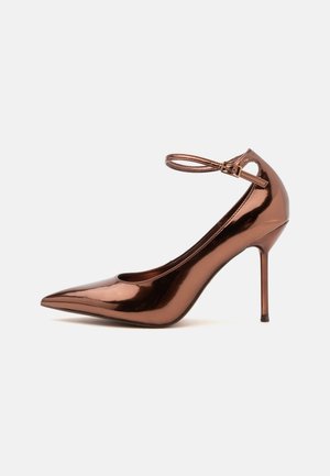 Chaussure à talon haut métallique bronze brillant avec bout pointu et fine bride de cheville avec boucle, sur un fond clair uni.
