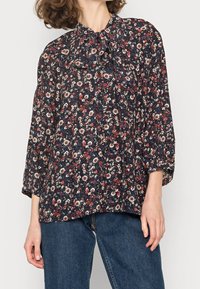 Femme portant un blouse floral ample avec un col à nœud, des manches longues, associée à un jean bleu sur un fond uni.