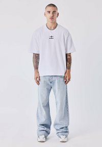 Witte katoenen t-shirt met korte mouwen en zwart logo, gecombineerd met lichtblauwe baggy jeans en witte sneakers met grijze accenten.