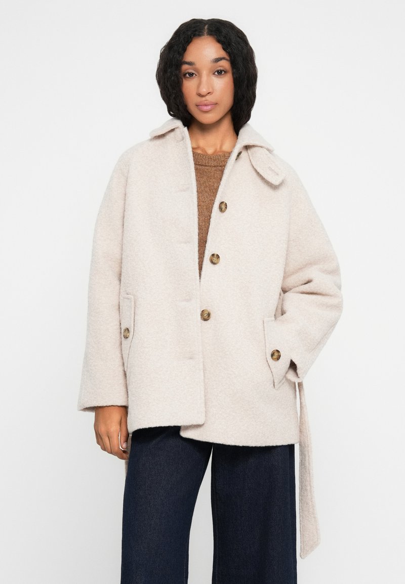 Cappotto in misto lana beige con ampio colletto, chiusura a bottoni singoli, due tasche anteriori e cintura in stoffa. Finitura testurizzata.