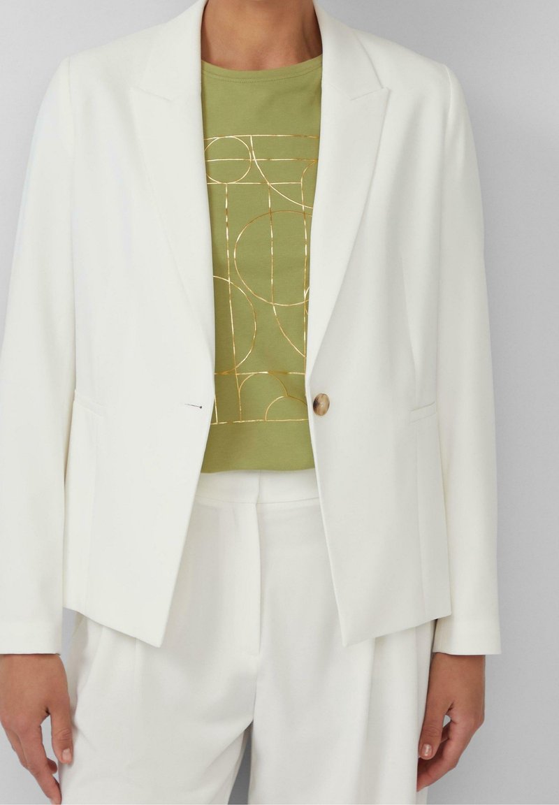 Blazer blanc à un bouton sur une chemise verte avec des lignes géométriques abstraites dorées, associé à un pantalon blanc assorti.