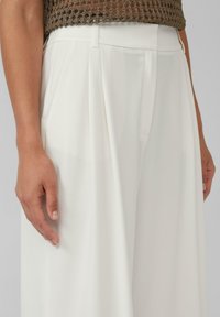Pantaloni bianchi plissettati realizzati in tessuto liscio, con una vita sagomata e tasche laterali. Una mano poggia sulla gamba dei pantaloni.