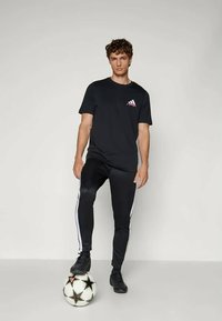 Black short-sleeve t-shirt with a red logo, paired with black athletic pants featuring white side stripes, standing on a black and white soccer ball.