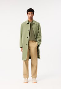 Ljusgrön regnkappa med svarta knappar, kombinerad med en olivgrön polo, beige byxor och vit-orange sneakers. Ren, minimal design.