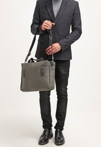 Szara skórzana torba listonoszka z dwoma przednimi kieszeniami, górnymi uchwytami i regulowanym paskiem, zestawiona z ciemnym marynarką i czarnymi dżinsami.