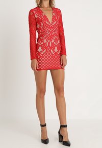 Robe en dentelle rouge à manches longues, décolleté profond en V, ornée de motifs floraux et de diamants, silhouette ajustée, longueur au-dessus du genou, associée à des talons noirs.