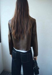 Brązowa skórzana kurtka z krótkim rękawem noszona na białym topie i ciemnych dżinsach. Czarna torebka trzymana w jednej ręce. Teksturowane włosy opadają luźno.