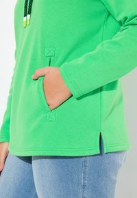 Felpa verde con tasca, spacchi laterali e cappuccio con stringa testurizzata. Il tessuto sembra morbido ed elastico. Abbinata a jeans azzurri chiari.