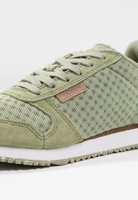 Baskets vert clair avec mesh texturé et panneaux en daim, lacets plats, étiquette logo marron, semelle intermédiaire blanche et semelle extérieure verte tachetée.