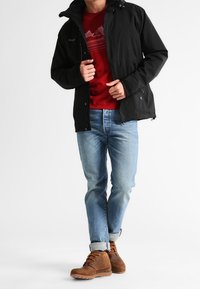 Czarna kurtka zapinana na zamek, czerwona koszulka z grafiką, jasnoniebieskie dżinsy i brązowe buty sznurowane. Kurtka ma kaptur i boczne kieszenie.