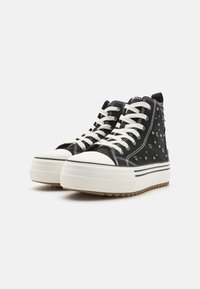 Zapatillas altas de lona negra con punteras de goma blanca y suelas de plataforma. Presentan tachuelas metálicas y cordones blancos en contraste.