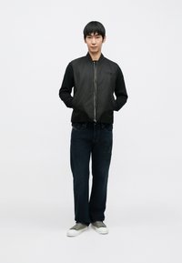 Crna bomber jakna jednostavnog dizajna, zatvaranje na patentni zatvarač; uparena s tamnoplavim trapericama s detaljima zakrpa i sivim tenisicama.