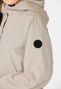 Veste beige avec capuche, en tissu texturé. Patch logo rond noir sur la manche gauche. Finition lisse, design décontracté.