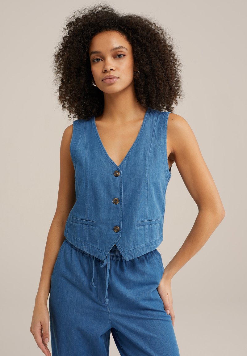 Modra denim brezrokavnica z V-izrezom, sprednjim zapiranjem na gumb in dvema žepoma. Rob je krajšan in ima obrobo z raztrganim robom.