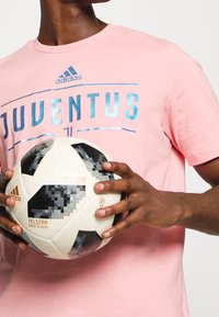 Růžové bavlněné tričko s nápisem "JUVENTUS" v reflexních modrých písmenkách; drží černobílý fotbalový míč s texturovaným povrchem.