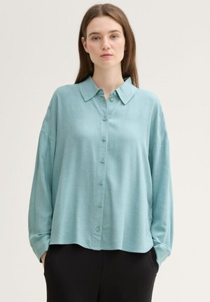 Blusa de manga larga, azul claro, con botones al frente, cuello tipo camisa y un corte relajado. Está hecha de una tela suave y texturizada.
