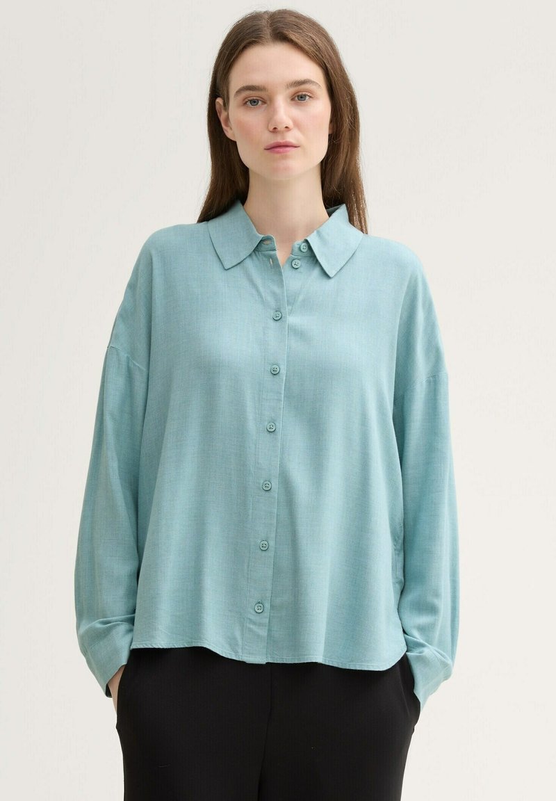Blusa de manga larga, azul claro, con botones al frente, cuello tipo camisa y un corte relajado. Está hecha de una tela suave y texturizada.