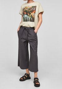 Beżowa koszulka z nadrukiem kolorowych liści, zestawiona z luźnymi, ciemnoszarymi spodniami 3/4 oraz czarnymi sandałami na paski.