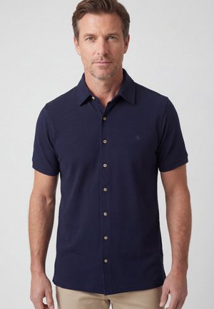 Mann trägt ein marineblaues kurzärmeliges Hemd mit Knöpfen und beige Hose, steht vor einem einfarbigen hellgrauen Hintergrund.