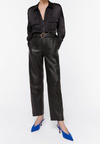 Camicia nera in satin con due tasche sul petto, abbinata a pantaloni in pelle nera a vita alta e décolleté blu a punta.