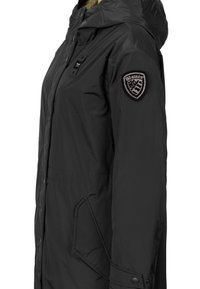 Sort isoleret jakke med hætte, der har en sidelomme med lynlås og et mærke på venstre arm. Glat tekstur og skræddersyet design.