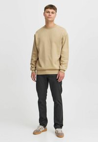 Beige crewneck sweatshirt, relaxed fit, gemaakt van zacht materiaal. Zwarte slim-fit broek, gecombineerd met grijze en witte sneakers. Eenvoudige, casual outfit.
