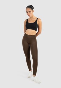 Leggings marroni a vita alta con una texture liscia, abbinati a un top corto nero. Modella in piedi contro uno sfondo neutro, indossando scarpe da ginnastica bianche.