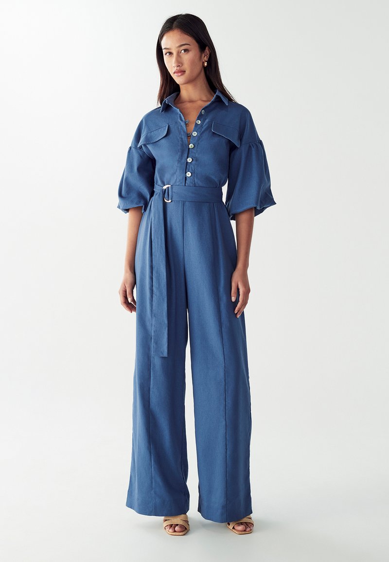 Combinaison en denim bleu avec un col, fermeture à boutons sur le devant, manches bouffantes, taille ceinturée et jambes larges. Texture lisse sur l'ensemble.
