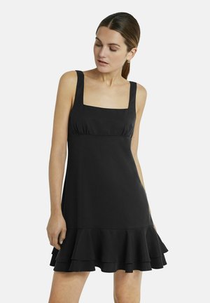 Frau mit braunen Haaren im tiefen Pferdeschwanz trägt ein ärmelloses schwarzes Kleid mit quadratischem Ausschnitt und abgestuften Volant-Saum, blickt nach unten.