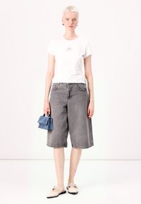 Biely tričko s potlačou, šedé denimové culottes a modrá zložená kabelka. Modelka má na sebe béžové ploché topánky. Svetlé pozadie.