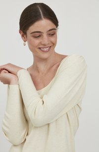 Hellbeige, locker sitzender Pullover mit langen Ärmeln, rundem Halsausschnitt und subtiler Ripptextur, getragen von einer Person mit verschränkten Armen.