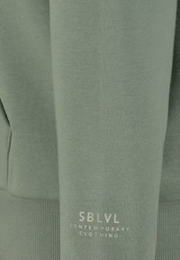 Groene sweatshirt van zachte stof met ribbelboorden en -onderkant. Heeft witte tekst "SBLVL CONTEMPORARY CLOTHING" nabij de onderkant.