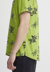 Camisa de manga corta en tejido verde brillante, con patrones de hojas de palma negras, detalles de botones y aberturas laterales para mayor transpirabilidad.