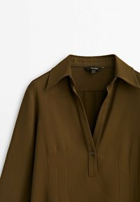 Blouse marron avec un col, patte de boutonnage à l'avant et détails de plis au niveau de la poitrine. Tissu lisse et léger.