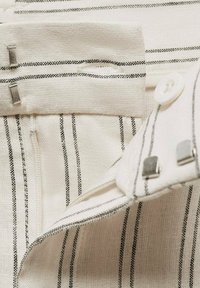 Witte stof met zwarte verticale strepen, voorzien van een knoopsluiting en metalen accenten. Gelaagde oppervlakte met gestikte randen.