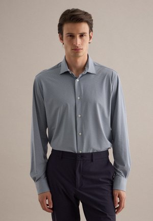 Mann steht vor neutralem Hintergrund, trägt ein hellblaues Hemd mit Knöpfen und dunkle Hose, schaut nach vorne mit neutralem Gesichtsausdruck.