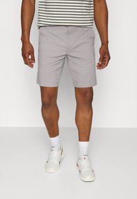 Un homme portant un short gris clair, des baskets Reebok blanches, des chaussettes blanches et une chemise rayée verte et blanche se tenant sur un fond blanc uni.