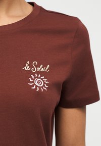 Καφέ μπλουζάκι με κείμενο «le Soleil» σε κρεμ χρώμα και ροζ γραφικό ήλιου στο αριστερό στήθος, φορεμένο από άτομο με ανοιχτό καφέ χρώμα δέρματος.