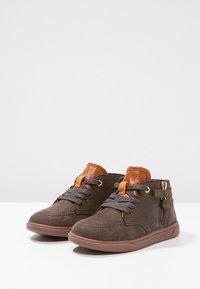 Zapatillas altas de ante marrón con cordones gris oscuro, acentos de lengüeta de piel color beige, cremallera lateral y suelas de goma sobre una superficie blanca.