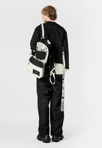 Vista trasera de una persona que lleva una mochila en blanco y negro, una camiseta de manga larga negra y pantalones negros con textura y detalle de etiqueta.