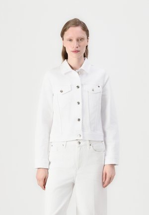 Kvinde iført en hvid knap-denimjakke og hvide bukser stående mod en ensfarvet hvid baggrund.