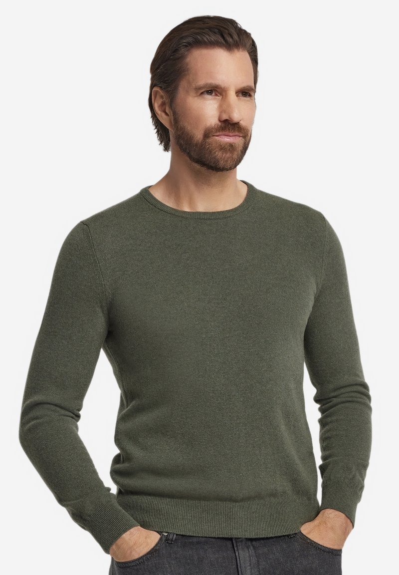 Mann mit braunen Haaren und Bart, der einen olivgrünen Pullover mit Rundhalsausschnitt und dunkle Jeans trägt, die Hände in den Taschen, schaut nach rechts.