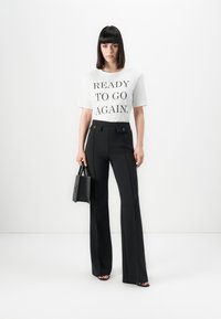 Woman standing in black high-waist pants, white T-shirt with "READY TO GO AGAIN" text, holding a black handbag, against a white background.
