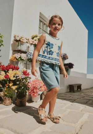 Mladá dívka v modrém květovaném topu a džínových šortkách kráčí po kamenné cestě vedle bílé zdi s barevnými květináči pod jasným slunečním svitem.