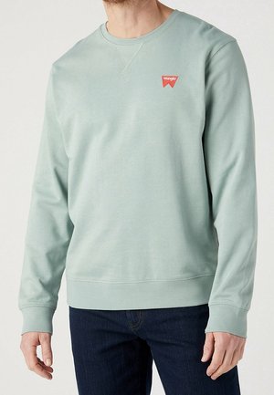 Mężczyzna ubrany w jasnozieloną, długorękawową bluzę z okrągłym dekoltem, z małym czerwonym logo Wrangler na piersi, w połączeniu z ciemnoniebieskimi dżinsami.