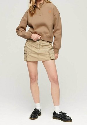 Woman wearing a brown pullover, beige cargo mini skirt, white socks, and black loafers standing against a plain background.