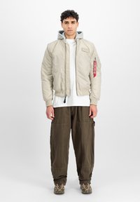 Light beige bomber jacket with grey hooded lining, zip front, ribbed cuffs, and patch detail. Worn over a white t-shirt and dark cargo pants.