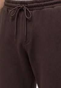 Gros plan d'une personne portant un pantalon de survêtement marron foncé avec une taille élastique et un cordon noué sur le devant.