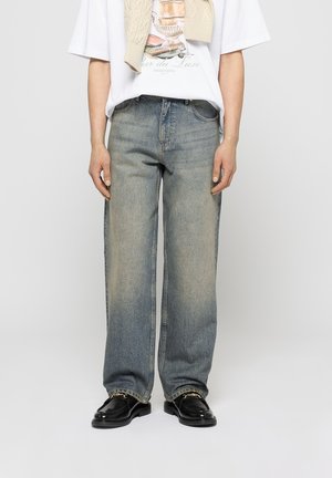 Homme portant un jean droit délavé, des mocassins noirs avec un détail de chaîne dorée, un t-shirt graphique blanc et un pull beige drapé sur les épaules.