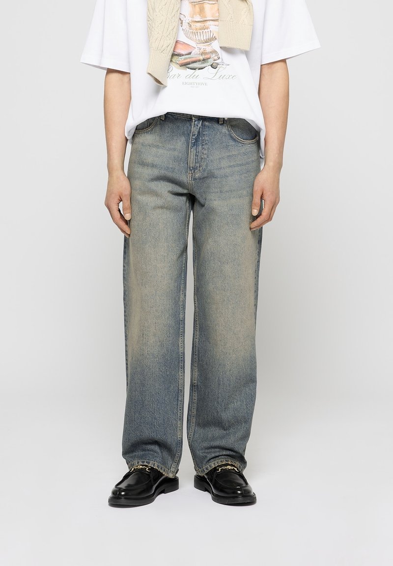 Homme portant un jean droit délavé, des mocassins noirs avec un détail de chaîne dorée, un t-shirt graphique blanc et un pull beige drapé sur les épaules.